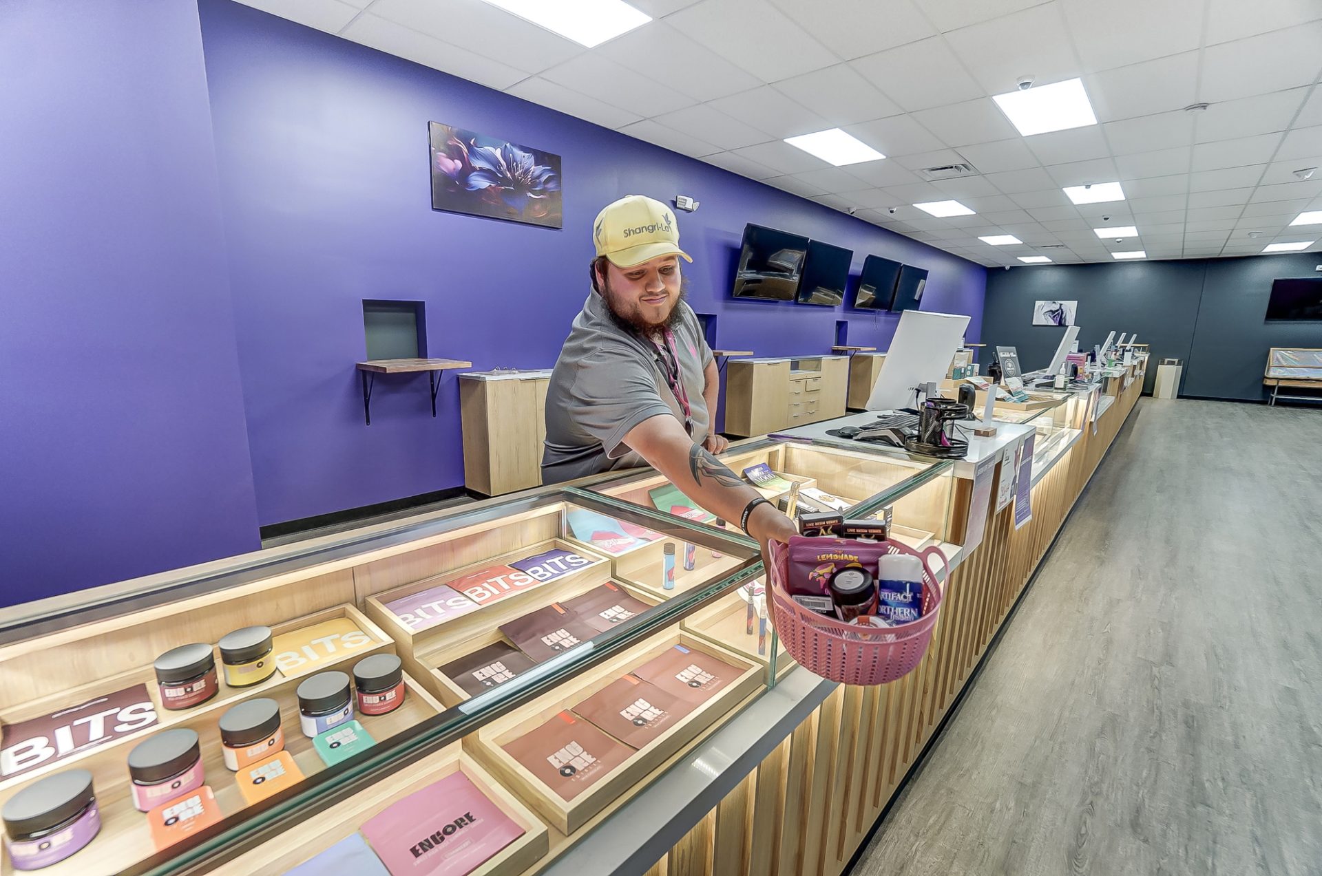A staff member hands over a pink basket of products across a modern retail counter with colorful packaged goods on display